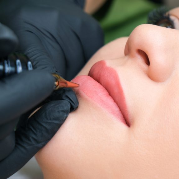 Young woman having permanent makeup on her lips at the beauticians salon. Natural green background